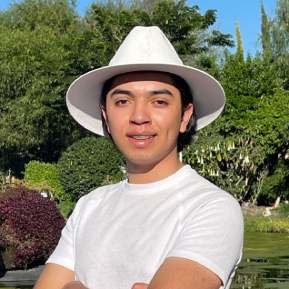 Retrato de Daniel, fondo con árboles y arbustos, viste una camiseta blanca con un sombrero blanco.